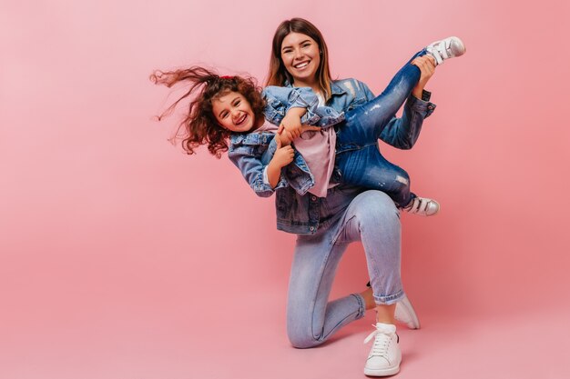 Mulher jovem sorridente brincando com a filha. Foto de estúdio de mãe feliz e criança pré-adolescente em trajes jeans.