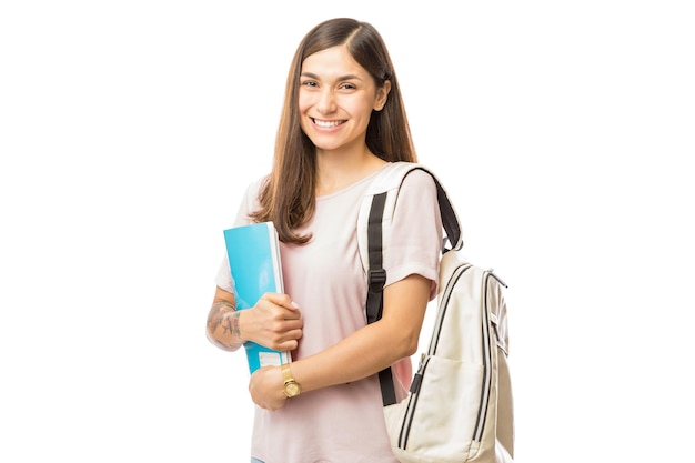 Mulher jovem sorridente com livros e mochila em pé sobre fundo branco