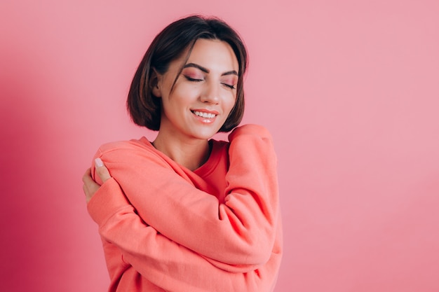 Mulher jovem sorridente de 20 anos em roupas casuais, isolada no retrato de estúdio de fundo rosa. conceito de amor. mantendo os olhos fechados de mãos cruzadas se abraçando