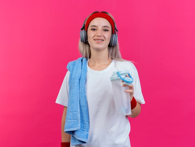 Mulher jovem sorridente e esportiva com aparelho nos fones de ouvido, bandana e pulseira segurando uma garrafa de água e uma toalha no ombro isolado na parede rosa