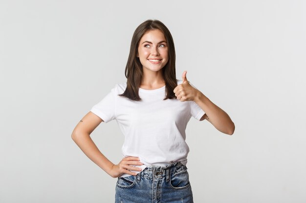 Mulher jovem sorridente e satisfeita mostrando o polegar para cima em aprovação, olhando no canto superior esquerdo