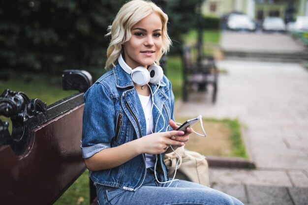 Mulher jovem sorridente feliz ouvindo música em fones de ouvido e usando o smartphone enquanto está sentado no banco da cidade