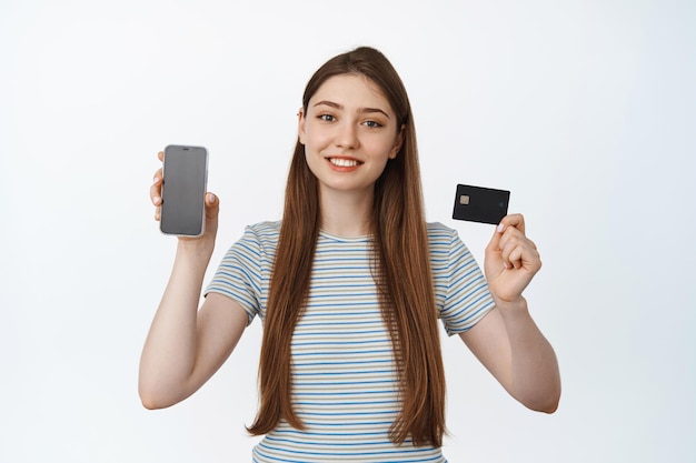 Mulher jovem sorridente mostrando a tela do telefone e cartão de crédito, mobile banking e conceito de compras, fundo branco