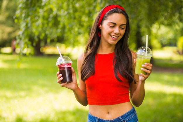 Mulher jovem sorridente segurando smoothies verdes e vermelhos em copo de plástico