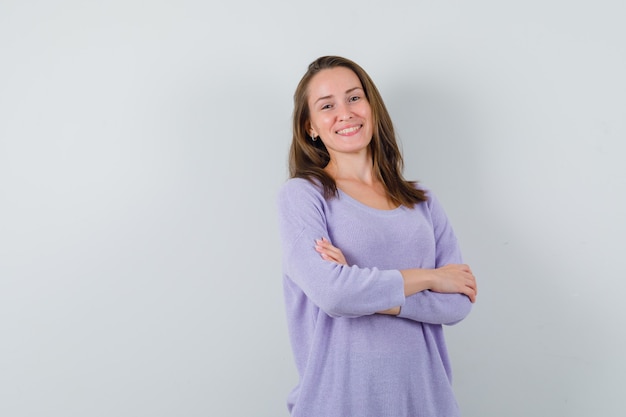 Mulher jovem sorrindo com os braços cruzados na blusa lilás e parecendo otimista