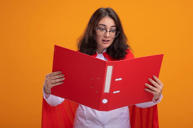 Foto grátis mulher jovem super-heroína impressionada com uma capa vermelha usando uniforme de médico e estetoscópio com óculos segurando e olhando para uma pasta isolada na parede laranja