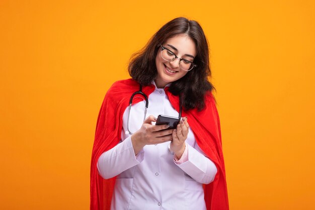 Mulher jovem super-heroína sorridente com capa vermelha, vestindo uniforme de médico e estetoscópio com óculos, usando seu telefone celular isolado na parede laranja com espaço de cópia