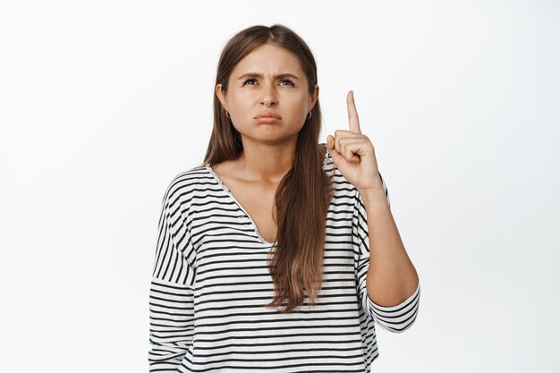 Mulher jovem suspeita, apontando o dedo para cima e olhando para o anúncio com olhos vesgos e expressão de rosto duvidoso, fundo branco.