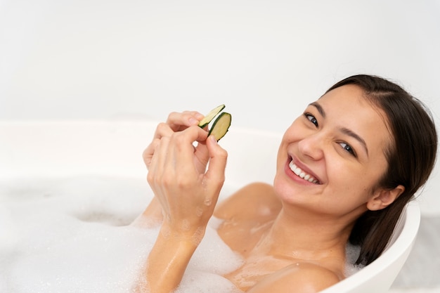 Mulher jovem tirando o pepino dos olhos e relaxando na banheira cheia de água e espuma