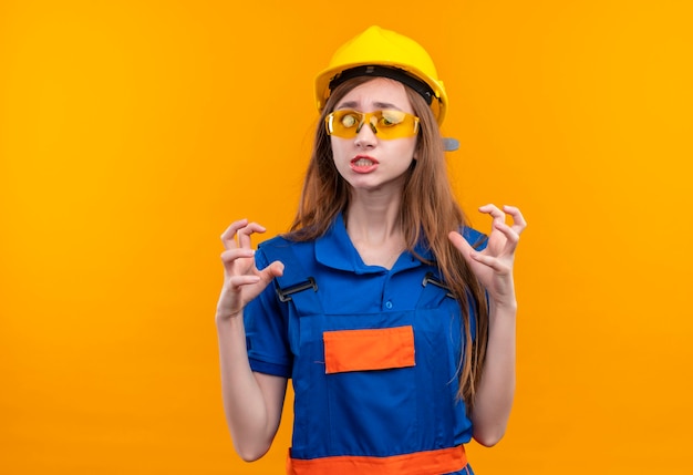 Mulher jovem, trabalhadora em uniforme de construção e capacete de segurança, sentindo ódio por alguém gesticulando com uma expressão agressiva em pé sobre a parede laranja