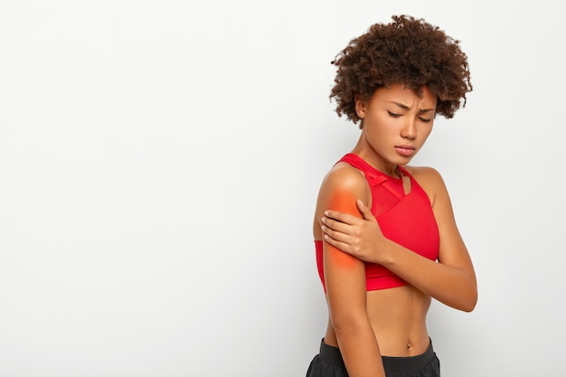 Mulher jovem triste sofre de sensação de dor no músculo do braço, fica de lado contra um fundo branco, ombro machucado durante a sessão de treino