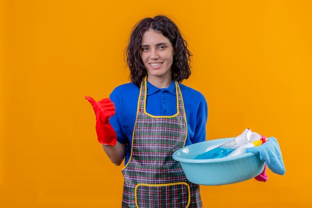 Mulher jovem usando avental e luvas de borracha segurando a bacia com ferramentas de limpeza, olhando para a câmera com um grande sorriso no rosto mostrando os polegares em pé sobre um fundo laranja