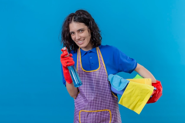 Foto grátis mulher jovem usando avental e luvas de borracha segurando a base com ferramentas de limpeza e spray de limpeza, olhando para a câmera, sorrindo alegremente em pé sobre um fundo azul