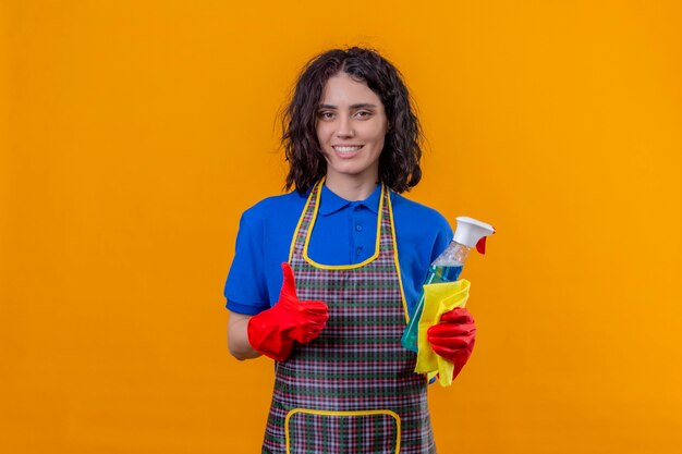 Mulher jovem usando avental e luvas de borracha segurando um spray de limpeza e um tapete, olhando para a câmera com um grande sorriso no rosto mostrando os polegares em pé sobre um fundo laranja