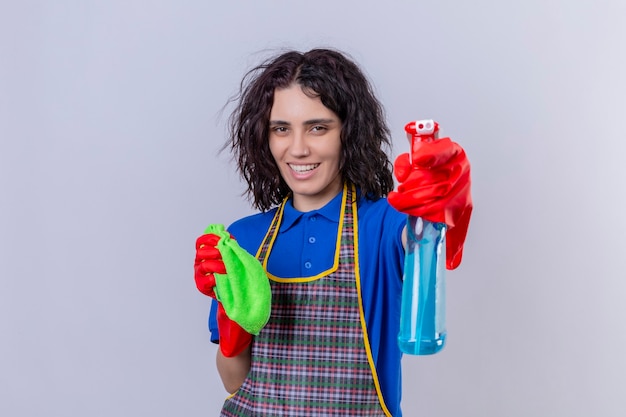 Foto grátis mulher jovem usando avental e luvas de borracha segurando um tapete e spray de limpeza se divertindo, sorrindo alegremente em pé sobre um fundo branco