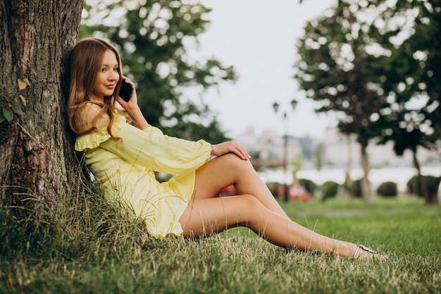 Mulher jovem usando o telefone e sentada sob uma árvore no parque