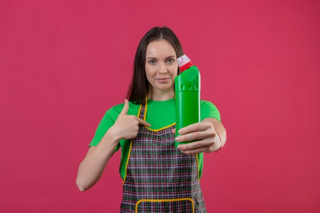 Mulher jovem vestida de uniforme segurando agente de limpeza e apontando o dedo para ela mesma na parede rosa isolada