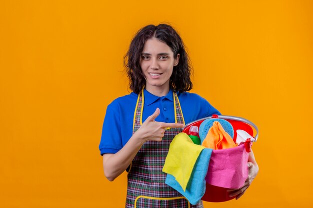 Mulher jovem vestindo avental segurando um balde com ferramentas de limpeza apontando com o dedo para ele e sorrindo em pé sobre um fundo laranja isolado