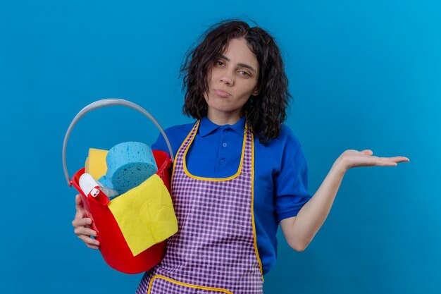 Mulher jovem vestindo avental segurando um balde com ferramentas de limpeza, parecendo confusa, sem resposta em pé sobre um fundo azul