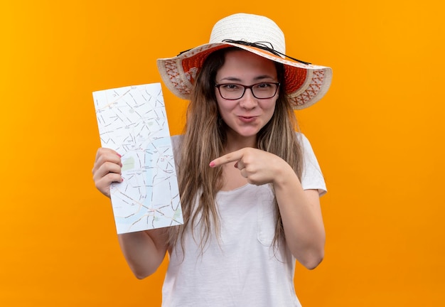 Foto grátis mulher jovem viajante com camiseta branca e chapéu de verão segurando um mapa apontando com o dedo para ele e sorrindo com uma cara feliz em pé sobre a parede laranja