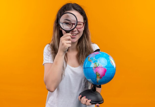 Mulher jovem viajante com camiseta branca segurando um globo, olhando pela lupa, parecendo surpresa e feliz em pé sobre a parede laranja