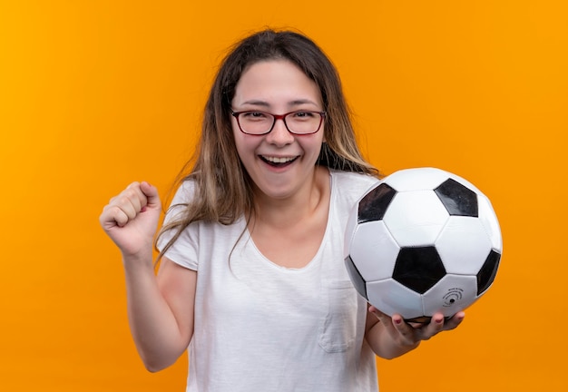 Mulher jovem viajante com camiseta branca segurando uma bola de futebol, parecendo animada e feliz com o punho cerrado em pé na parede laranja