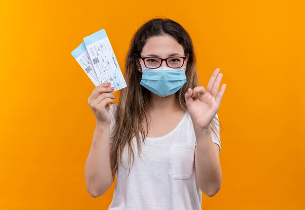 Mulher jovem viajante com camiseta branca usando máscara facial protetora segurando passagens aéreas sorrindo fazendo sinal de ok em pé sobre a parede laranja