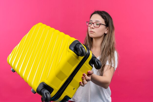 Mulher jovem viajante com uma camiseta branca segurando uma mala de viagem olhando para o lado com uma cara séria em pé sobre a parede rosa