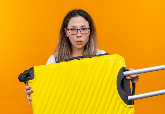 Mulher jovem viajante com uma camiseta branca segurando uma mala, parecendo confusa e muito ansiosa em pé sobre a parede laranja