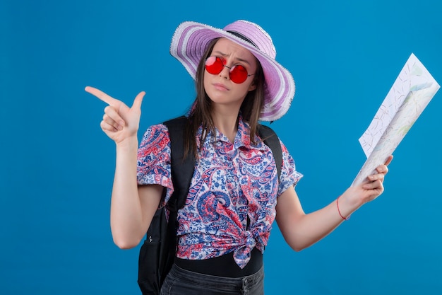 Mulher jovem viajante de chapéu de verão usando óculos de sol vermelhos, segurando o mapa olhando de lado com expressão pensativa, apontando com o dedo para o lado pensando tendo dúvidas sobre parede azul