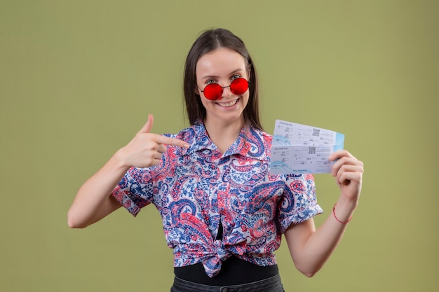 Foto grátis mulher jovem viajante de óculos vermelhos segurando bilhetes apontando com o dedo para eles sorrindo com cara feliz sobre parede verde