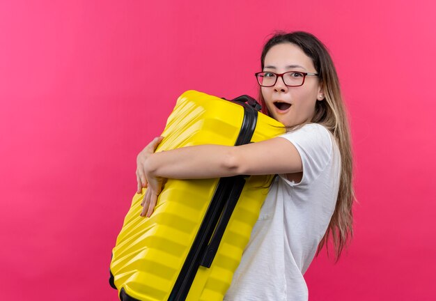 Mulher jovem viajante em uma camiseta branca abraçando sua mala de viagem, parecendo espantada e surpresa em pé sobre uma parede rosa