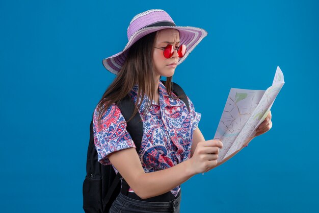 Mulher jovem viajante no chapéu do verão de óculos vermelhos, segurando o mapa olhando para ele com o rosto carrancudo sobre parede azul