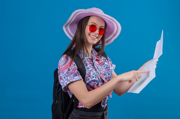 Mulher jovem viajante no chapéu do verão usando óculos de sol vermelhos, segurando o mapa apontando com o dedo para ele sorrindo alegremente sobre parede azul