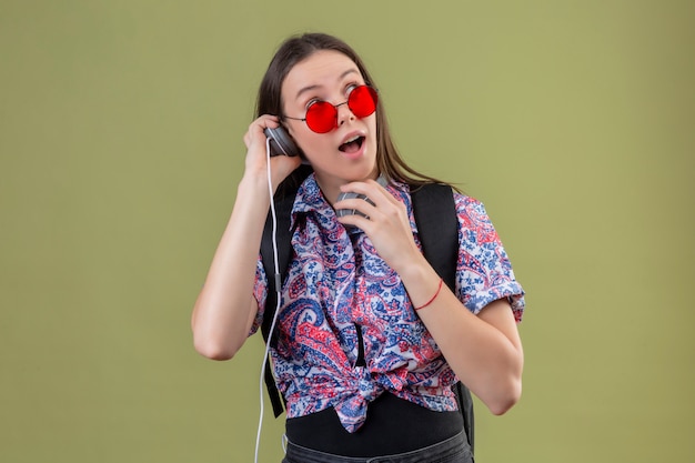 Mulher jovem viajante usando óculos de sol vermelhos e com mochila, ouvindo música usando fones de ouvido, olhando surpreso e feliz ao longo da parede verde