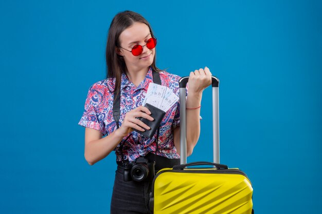 Mulher jovem viajante usando óculos escuros vermelhos em pé com uma mala amarela segurando o passaporte e as passagens, olhando de lado positiva e feliz sobre o fundo azul