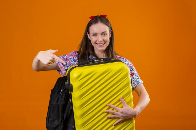 Mulher jovem viajante usando óculos escuros vermelhos na cabeça, com uma mochila segurando a mala, olhando para uma câmera e apontando com o dedo para a mala, sorrindo com uma carinha feliz sobre fundo laranja