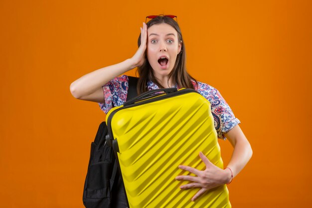 Mulher jovem viajante usando óculos escuros vermelhos na cabeça, em pé com uma mochila segurando uma mala, parecendo surpresa e maravilhada com a mão na cabeça e boca bem aberta e olhos sobre fundo laranja