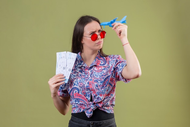 Mulher jovem viajante usando óculos escuros vermelhos segurando bilhetes e avião de brinquedo, olhando de lado com expressão pensativa com o rosto carrancudo sobre parede verde
