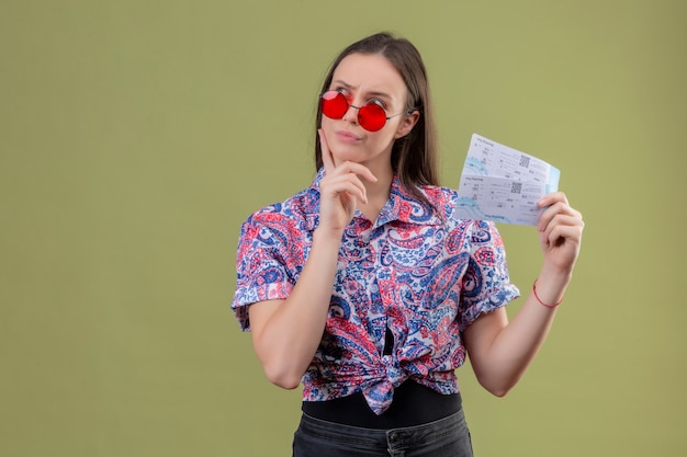 Mulher jovem viajante usando óculos escuros vermelhos segurando bilhetes olhando de lado com expressão pensativa descontente sobre parede verde