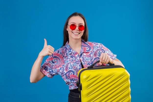 Mulher jovem viajante usando óculos escuros vermelhos segurando uma mala amarela, olhando para a câmera, sorrindo alegremente, mostrando os polegares em pé sobre um fundo azul