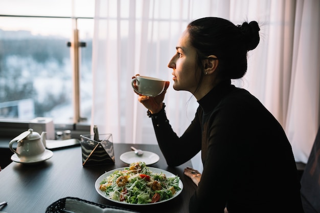 Foto grátis mulher jovem, xícara segurando, de, bebida, tabela, com, salada, perto, janela