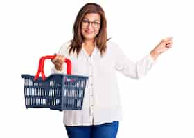 Foto grátis mulher latina de meia-idade segurando cesta de compras de supermercado sorrindo feliz apontando com a mão e o dedo para o lado