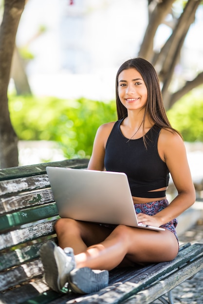 Mulher latina satisfeita sentada no banco do parque e usando o laptop