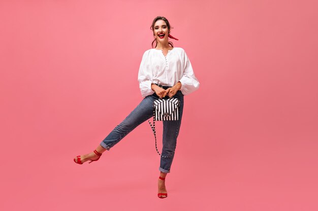 Mulher legal em jeans e blusa segurando a bolsa no fundo rosa. Menina elegante com batom vermelho e bandagem no cabelo está se divertindo.