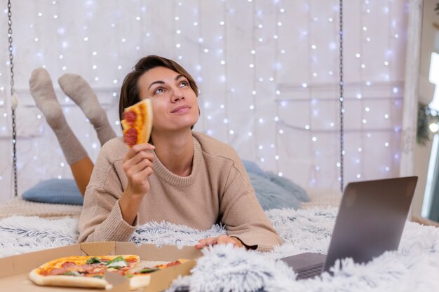 Mulher leste fast-food de entrega na cama no quarto em casa. Mulher sozinha apreciando comida gordurosa, pizza