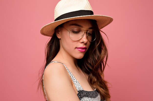 Mulher linda e atraente com cabelo ondulado, chapéu e óculos com lábios rosados posando sobre uma parede rosa