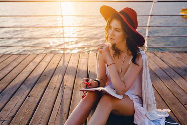 Mulher linda em um vestido branco sentada à beira-mar no nascer do sol pensando e fazendo anotações no livro diário