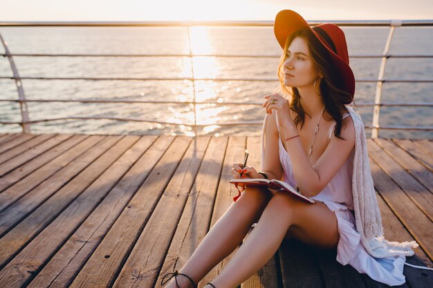 Mulher linda em um vestido branco sentada à beira-mar no nascer do sol pensando e fazendo anotações no livro diário