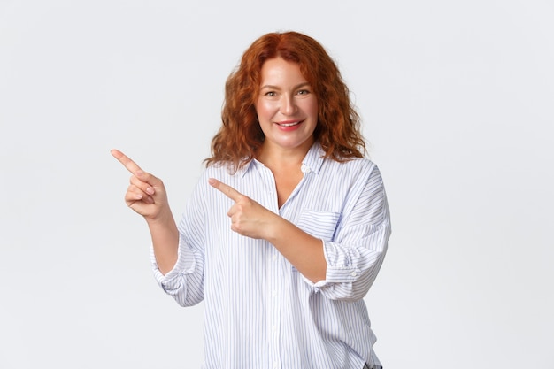 Mulher linda ruiva de meia-idade sorridente mostrando o anúncio, apontando o canto superior esquerdo dos dedos. Senhora alegre com cabelo ruivo demontrate bandeira de produto sobre fundo branco.